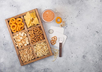 Glass of craft lager beer and opener with box of snacks on light background. Pretzel,salty potato sticks, peanuts, onion rings with nachos in vintage box with openers and beer mats. Space for text