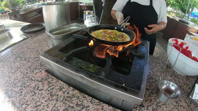 The cook prepares breakfast in a pan over an open fire. A female Turkish hotel cook cooks an omelet with bacon and vegetables over an open fire.