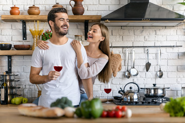 Beautiful laughing young couple drinking wine and cooking salad while standing in the kitchen