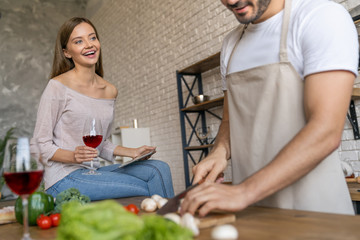 Beautiful young woman with glass of wine using a digital tablet and smiling while her man cooking in kitchen at home