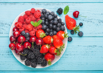 Fresh organic summer berries mix in white plate on blue wooden table background. Raspberries, strawberries, blueberries, blackberries and cherries.