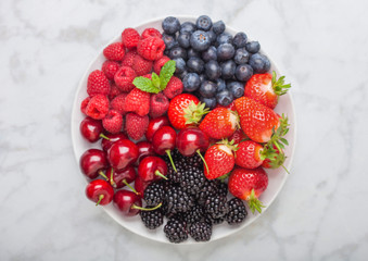 Fresh organic summer berries mix in white plate on marble background. Raspberries, strawberries, blueberries, blackberries and cherries.