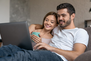 Smiling young couple relaxing on a couch and drinking tea at home while using laptop computer