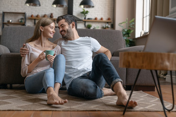 Young happy couple sitting on the floor at home and looking on each other while watching movie on laptop