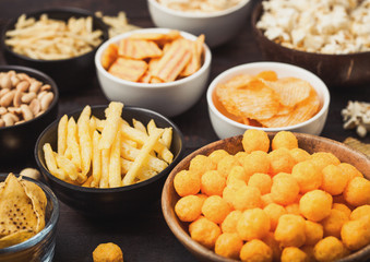 All classic potato snacks with peanuts, popcorn and onion rings and salted pretzels in bowl plates on wooden background. Twirls with sticks and potato chips and crisps with nachos and cheese balls.