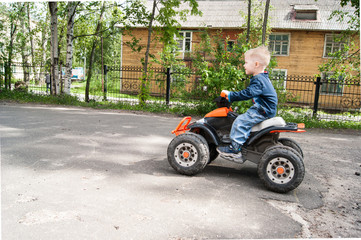 A fair-haired boy of three years in a leisure park rides along a track on a children's orange electro-quad