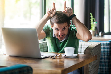 Young angry businessman in green t-shirt sitting and looking at camera with crazy face and cow horns gesture on his head. business and freelancing concept. indoor shot near big window at daytime.