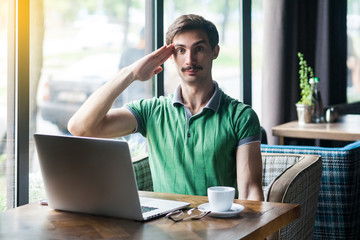 Yes sir! Young serious businessman in green t-shirt sitting and looking at camera with salute gesture and ready to complete the mission. business concept. indoor shot near big window at daytime.
