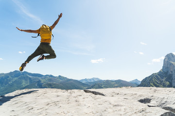 Young Man Jumping on Top of a Mountain Wearing Yellow Backpack. - obrazy, fototapety, plakaty
