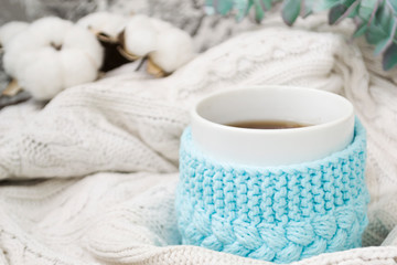 White mug with tea in a knitted blue frame and wrapped in a white knitted plaid. In the background, sprigs of eucalyptus and cotton. Breakfast in bed. Cozy.