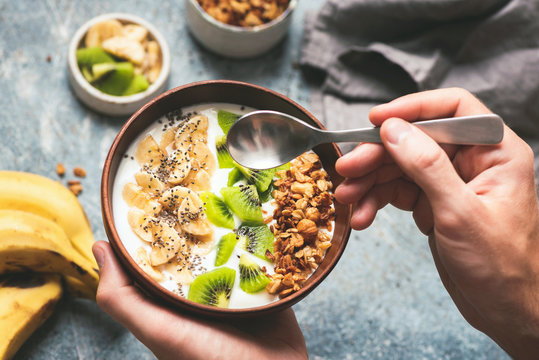 Eating healthy breakfast yogurt bowl with granola and fruits kiwi, banana, coconut. Male hands holding coconut bowl with yogurt and fruits
