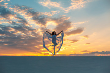 A girl in a fly white dress dances and poses in the sand desert at sunset