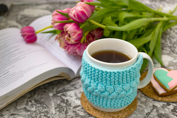 White cup of tea in a knitted blue case with an open book and with pink flowers tulips and sweets, with gingerbread on a textural table. Spring, winter concept