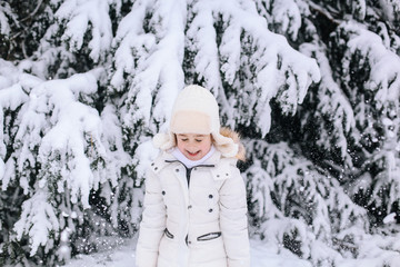 Fototapeta na wymiar Portrait of little girl in winter. Toddler playing with snow in park. Child in white warm winter clothes and knit hat looking at the falling snow In winter forest. Snowfall. Winter activities for kids
