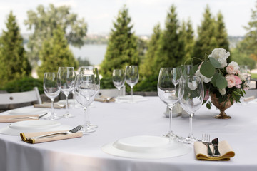 Wedding table setting decorated with fresh flowers in a brass vase. Wedding floristry. Banquet table for guests outdoors with a view of green nature. Bouquet with roses, eustoma and eucalyptus leaves