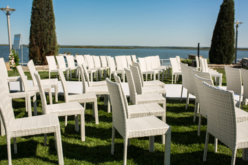 Beautiful outgoing wedding set up.Romantic wedding ceremony , wedding outdoor on the lawn water view. Wedding decor. White wooden chairs on a green lawn. White armchairs for guests