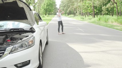 Female driver calling tow truck service after her car broke on countryside road. Selective focus on broken car with open hood, woman calling tow truck service on background