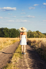 Beautiful young woman in a white dress and hat walks along the road between two fields with wheat. Concept of outdoor recreation, a trip to the village, vacation