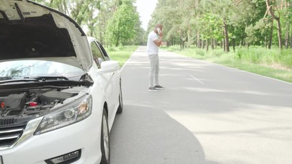 Man calling tow truck service on countryside road after his car broke down. Car with open hood on foreground, male driver calling his mechanic for help. Car workshop, repair concept