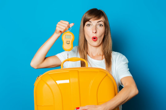 Beautiful young woman with a surprised face weighs a crowded yellow plastic suitcase on a blue background. Concept baggage, excess baggage, baggage weight
