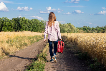 Beautiful young woman with a suitcase is walking along a road between wheat fields in the background. Travel concept, vacation