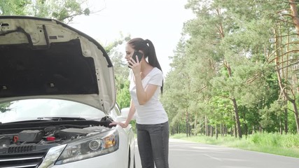 Beautiful woman looking under the hood of her broken car on countryside road. Young female driver examining machinery of her car, waiting for tow truck service help on the road