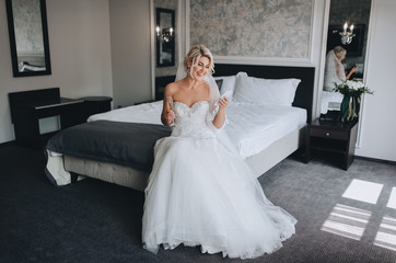 A beautiful, smiling blonde bride in a white dress is sitting on the bed and looking at the phone,...