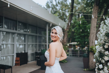 Beautiful blonde bride stands and smiles in a restaurant against the background of garlands and light bulbs. Wedding portrait of a cute girl close-up in a white dress. Concept and photography.