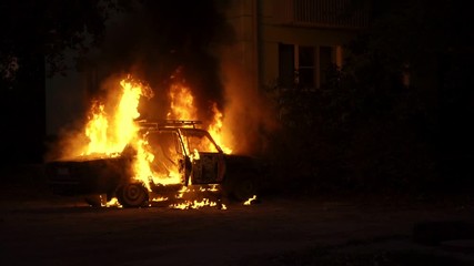 Burning car close to a house in the city of slow motion. Theme of criminal revenge and set fire to someone else car. Automobile engulfed in flames of fire. Trees close to burning sedan.