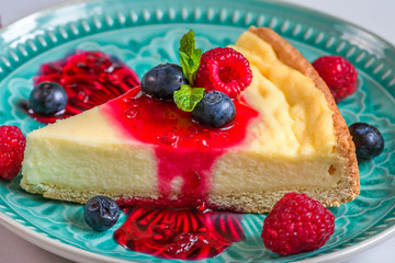slice of cheesecake with fresh raspberries, blueberries, jam and mint in a blue plate on marble background. close up
