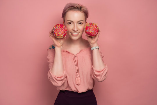 A girl with short pink hair, holds donuts in her face, smiling at the camera, on a pink background.