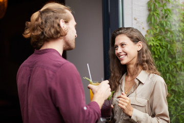 Waist up portrait of smiling young woman talking to man outdoors standing by cafe door, both holding cold refreshing drinks, copy space