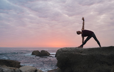 Man doing the triangle pose by the ocean at dusk
