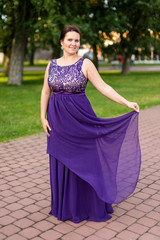 Smiling Caucasian brunette woman is touching hem of purple floor-length dress in park.