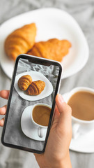 Fototapeta na wymiar Female hand with mobile phone taking picture of tasty breakfast coffee and croissants in bed, closeup