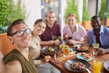 Multi-ethnic group of people posing for selfie photo while enjoying party dinner in cafe together, focus on smiling man in holding camera in foreground, copy space