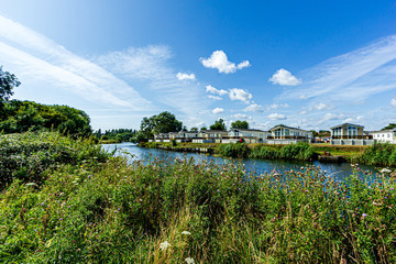 A trailer park along a river bank with wild green vegetations and trees on the foreground under a majestic blue sky and some white clouds