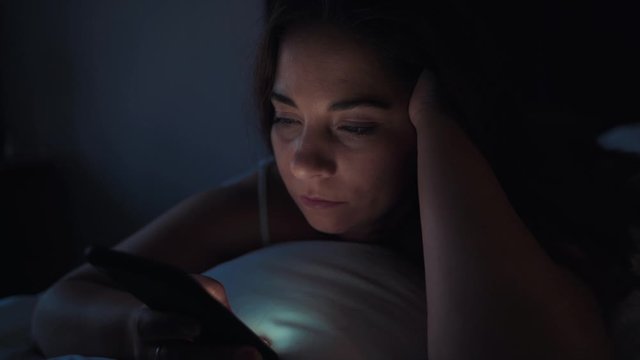 Young woman using smartphone lying in bed instead of sleep at night. Insomnia concept