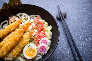 Fototapeta na wymiar Close up of bowl of gourmet Japanese udon noodle soup with colorful toppings of pickled ginger, boiled egg, swirly pattern narutomaki, and breaded prawns