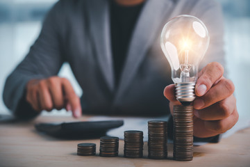 businessman hand holding lightbulb with coins stacking on desk.saving energy and money concept.