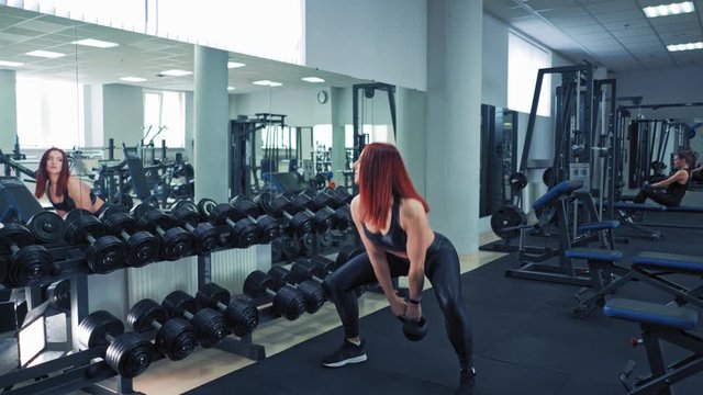 Sportswoman in black top and leggings swinging hard kettlebell in fitness club. Female athlete doing workout and looking at the mirror near rows of dumbbells.
