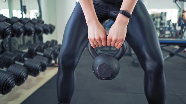 Sporty female lifting kettlebell. Athlete woman wearing black leggings and trainers training with hard weight in sports club. Close-up.