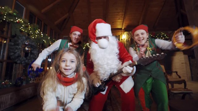 Happy Santa, elves and girl with musical instruments dancing and looking to camera. Funny Christmas atmosphere in Santa's residence.