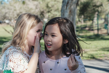 multiethnic young little girls playing kids Chinese whispering  in the park, best friends and...