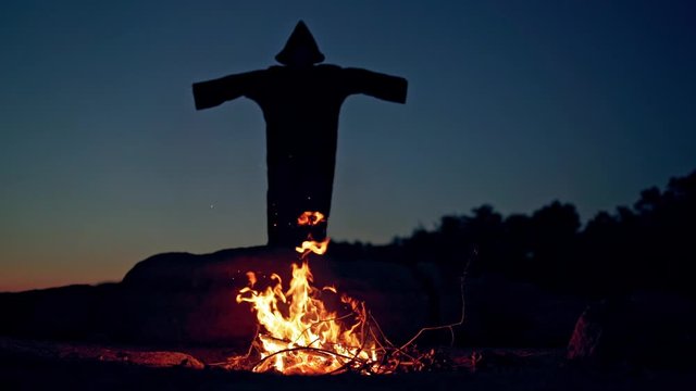 Spooky nightmare figure with long hood is turning. Small campfire and black silhouette of a witch at night.