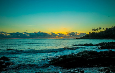 Sunrise over the ocean in Itacaré, Bahia, with silhouettes of the coastal landscape and coconut trees against a vibrant orange-to-blue sky, creating a serene tropical scene.
