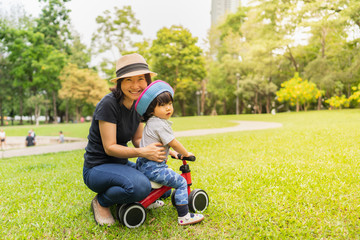 Young Asian mother smiling toward to camera with her toddler while learn how to ride a bike helped and protected by her mother in the autumn color season park, concept outdoor family activity time.
