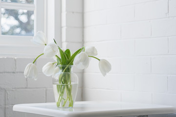 Close up of white tulips in glass vase on table against painted brick wall and window (selective focus)