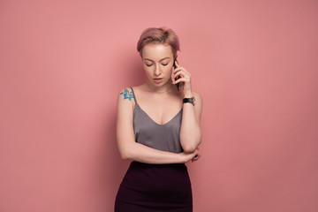 A girl with short pink hair and a tattoo in a gray top and speaks on the phone, looking down, standing on a pink background.