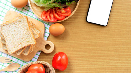 Homemade sandwich breakfast preparing. Whole wheat bread is stacked on a wooden cutting board. Smart phone, slice tomatoes, ham, eggs and lettuce on wooden plate. Cooking ingredient. Food background.